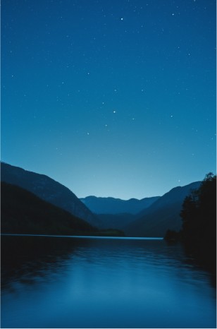 A tranquil nighttime scene features a starry sky, mountainous terrain, and still water, conveying serenity and vastness.