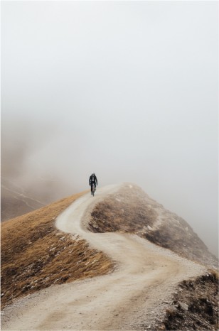 A solitary figure walks on a winding path along a ridge, surrounded by misty white fog, evoking a sense of contemplative isolation.